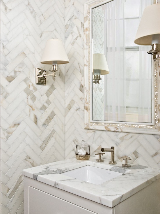 a small yet luxurious marble powder room with chevron clad walls and a chic mirror plus wall lamps