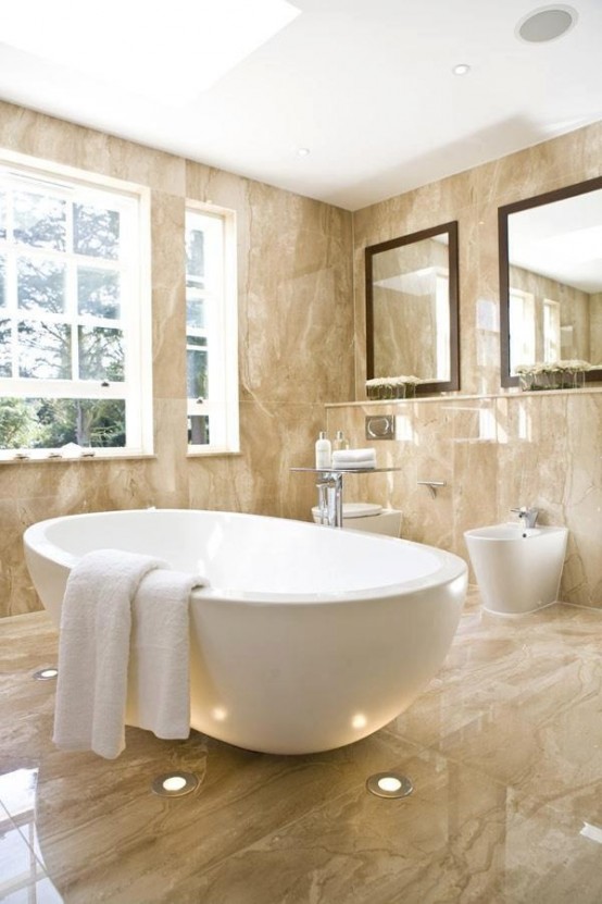 a beige bathroom fully clad with marble, with an oval tub, large mirrors and a window for more light