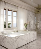 a luxurious white marble bathroom with a large window with curtains, a tub clad with marble and a shower space