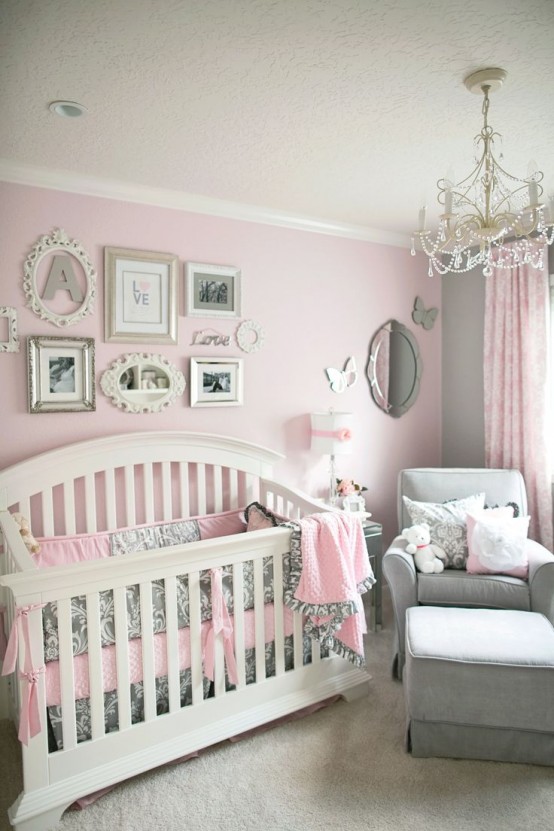 a refined vintage nursery with pink walls, a white crib, a grey chair and a footrest, a crystal chandelier and a gallery wall