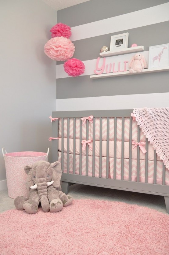 a pink and grey nursery with a striped accent wall, floating shelves, a grey crib with pink bedding and a pink rug, some toys