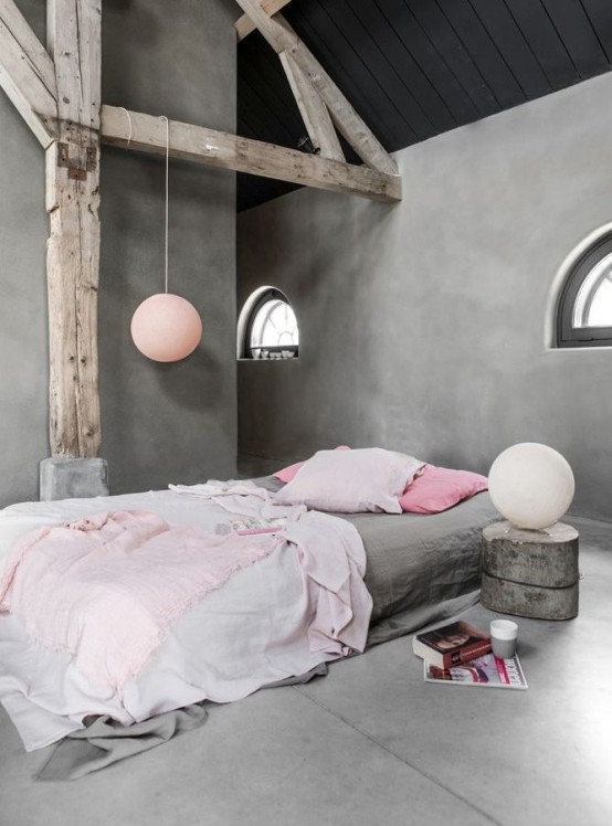 a wabi-sabi bedroom with concrete walls and a floor, a low bed with grey and pink bedding, wooden pillars and beams, some pendant lamps