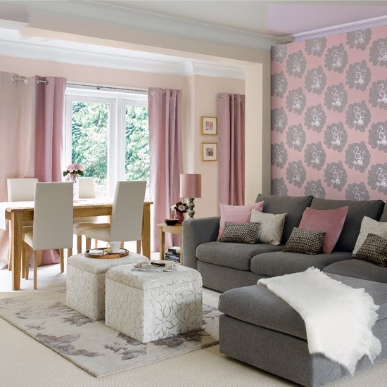 a welcoming living room with pink walls and an accent pink wallpaper wall, a low grey sofa, a stained table and white chairs and some creamy poufs