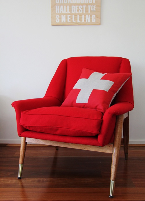 a catchy and bright mid century modern chair with a stained frame and legs and a red seat with a red cushion and pillow is comfy and bright