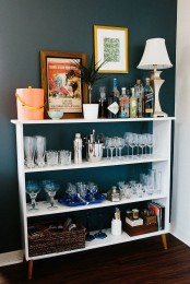 an open home bar made of a dresser with lots of glasses, bottles and other stuff you may need plus a lamp
