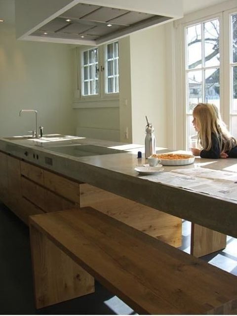 a minimalist kitchen with an oversized sleek wooden kitchen island and a concrete countertop plus wooden benches