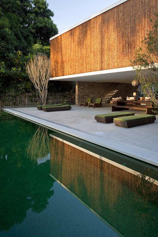 a minimalist terrace over the pool, with a concrete floor, wooden loungers with green cushions and pillows, potted trees and some more furniture in the open living room