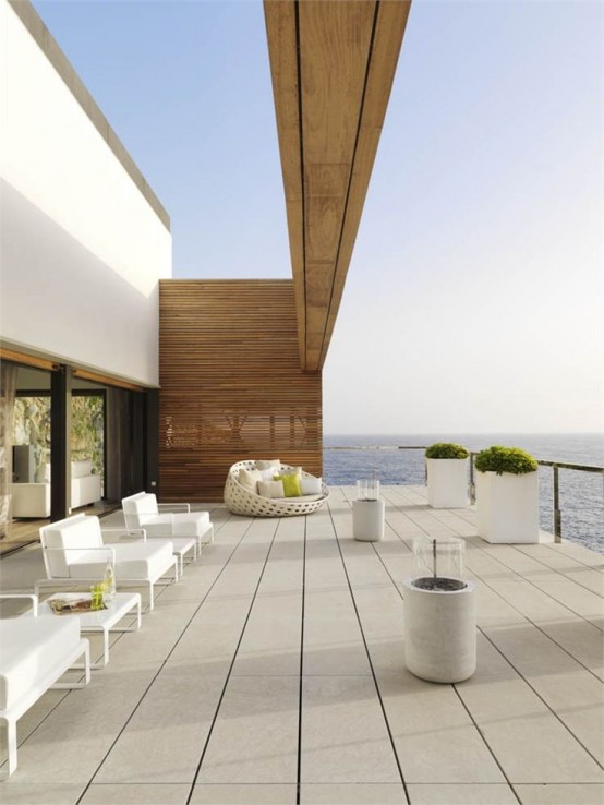 a minimalist sea view terrace with a planked floor, white outdoor furniture, a rounded chair with pillows, potted plants and some lamps plus a gorgeous view