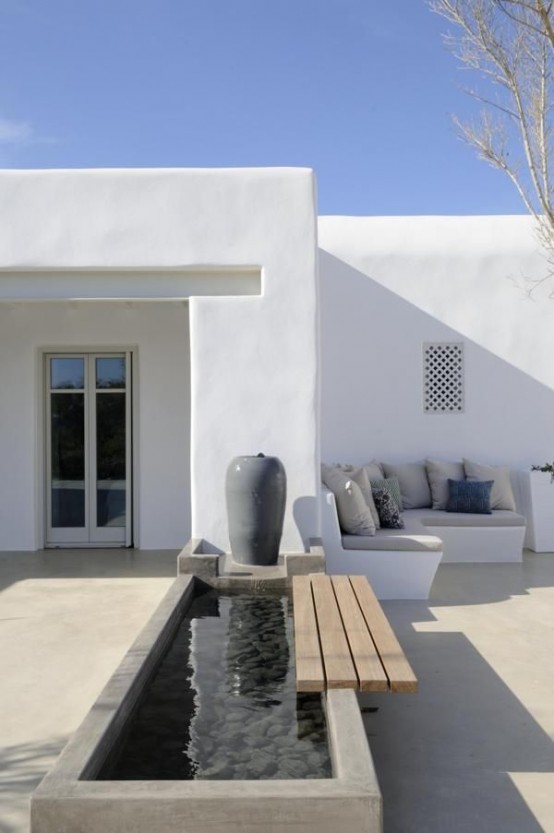 an ultra-minimalist outdoor space with a concrete floor and a concrete mini pond with pebbles, a large vase and a built-in bench with pillows