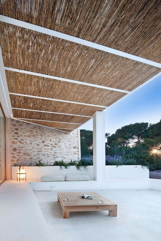 a minimalist terrace with a concrete floor and built-in benches, a low wooden coffee table, some pillows and a lantern plus a view of the garden