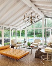 a modern sunroom with glazed walls, a taupe sofa, printed chairs, a coffee table, an orange leather daybed and a chic vintage chandelier