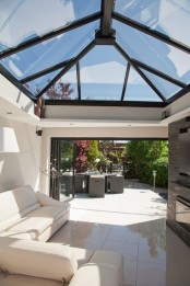 a minimal modern sunroom with a glass roof and a white leather sofa, a TV and a built-in fireplace plus a folding glass wall