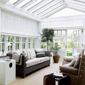 a modern farmhouse sunroom with a dark sofa with pillows, a couple of tan chairs, side tables and potted greenery, glazed walls and a glass roof