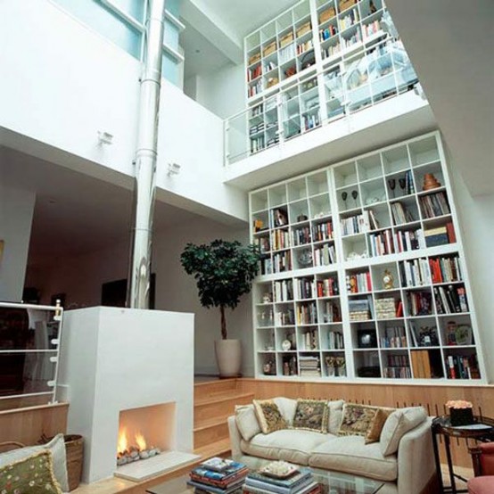 a double-height white library with lots of bookshelves, a sleek white fireplace and some neutral sitting furnitureplus skylights over it