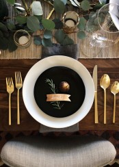 a modern Thanksgiving tablescape with a woven table runner, greenery, gold glitter, gold cutlery and black and white plates