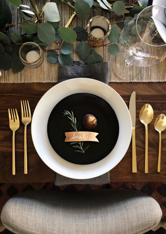a modern Thanksgiving tablescape with a woven table runner, greenery, gold glitter, gold cutlery and black and white plates
