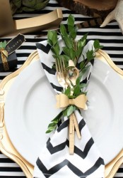 a modern Thanksgiving tablescape with a striped tablecloth and a chevron napkin, a gold chargers and gold cutlery, greenery attached to the napkin