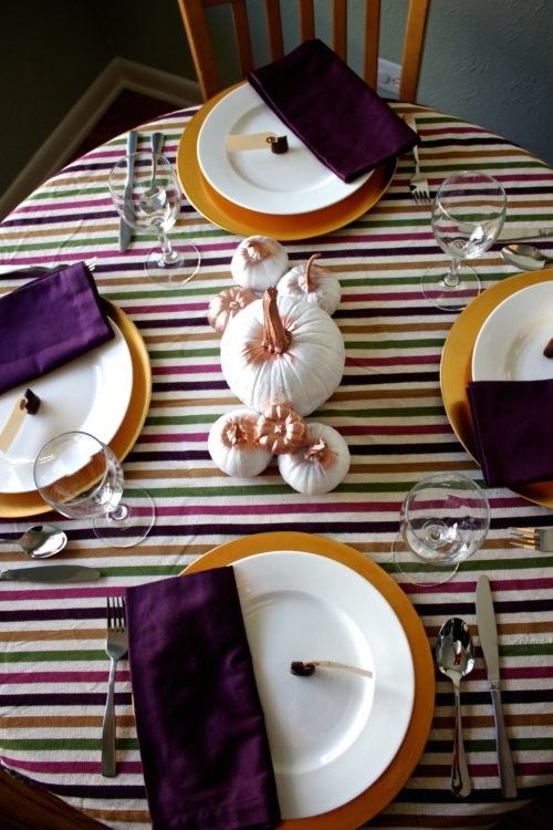 a modenr colorful Thanksgiving tablescape with a striped tablecloth, pumpkins, gilded chargers, deep purple napkins is a very chic one