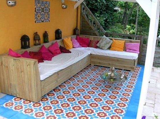 a bold outdoor space with an orange accent wall, a corner sofa with colorful pillows, a colorful Moroccan tile floor and Moroccan lanterns plus greenery around