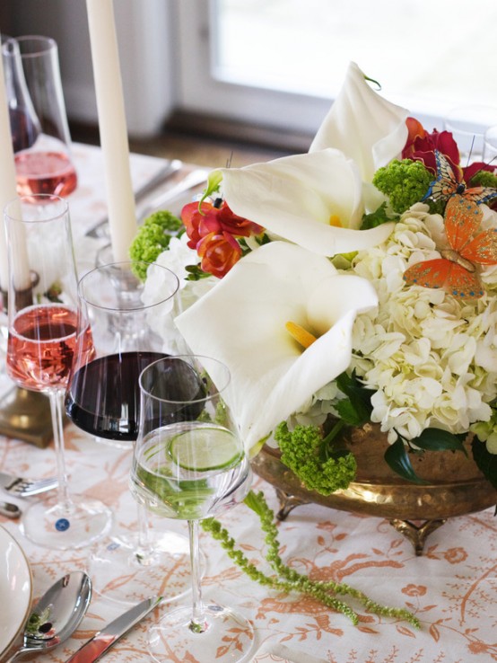 a delicate Mother's Day table setting with a floral tablecloth, white and bright blooms and greenery, tall and thin candles for more elegance