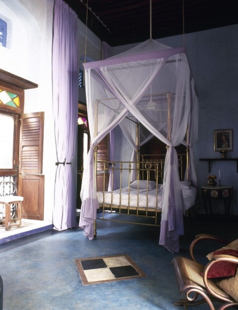 a Moroccan bedroom with a gold bed and lavender canopy, shutters and a mosaic balcony entrance, carved wooden furniture