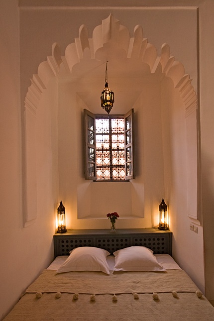 a small and neutral Moroccan bedroom in an alcove with a mosaic window, a storage headboard and lanterns