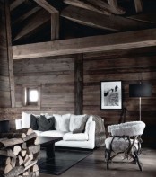 a modern chalet living room clad with wood, with a firewood storage, a white sofa and a chair and artwork is moody yet cozy