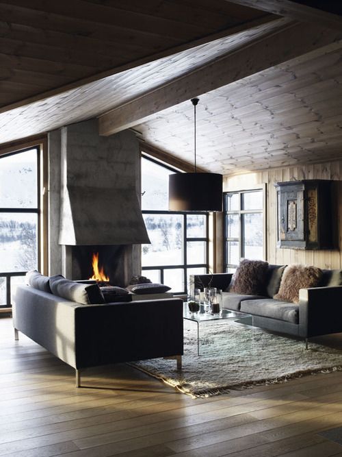 a restraint contemporary chalet living room with a glazed wall for the views, a hearth, grey sofas and an acrylic coffee table plus a cool chandelier