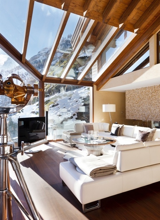 a fantastic contemporary chalet living room with glazed walls and a roof, white seating furniture, a TV, a chic round glass table and some cool lamps
