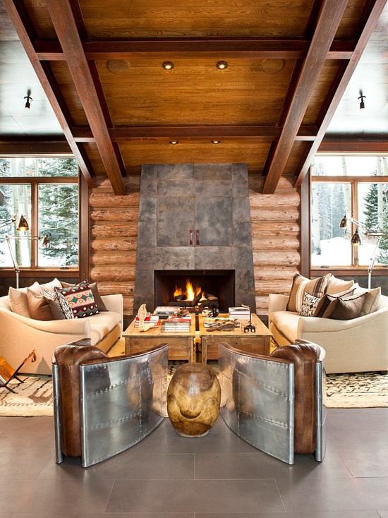 a cozy boho chalet living room with large windows and skylights, an aged metal clad fireplace, neutral sofas, wooden tables and metal and leather chairs