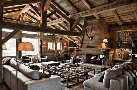 a modern chalet living room with multiple beams and pillars, a fireplace clad with wood, grey seating furniture, large coffee tables of wood and glass