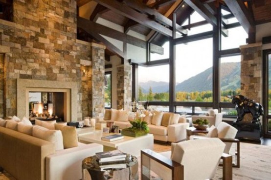 a neutral chalet living room with multiple black wooden beams, a glazed wall for a view, a large stone clad fireplace and creamy seating furniture