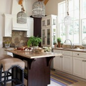 a neutral farmhouse kitchen with a deep purple kitchen island, stone countertops and a backsplash, crystal pendant lamps and greenery