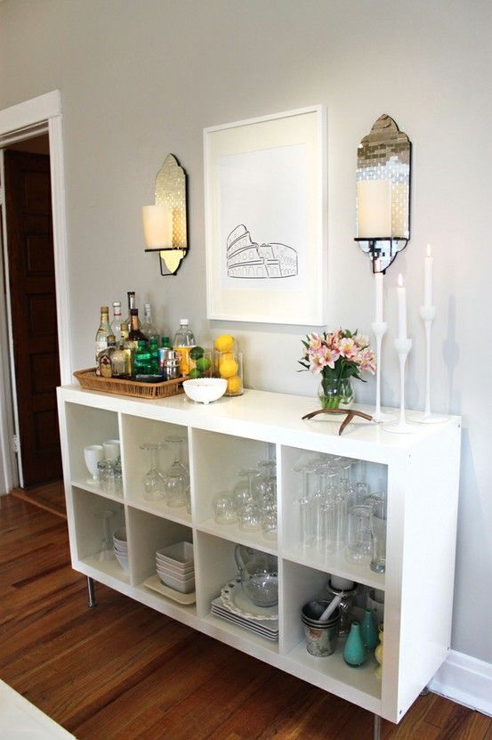 an IKEA Kallax piece turned into a home bar, with glass and bottle storage, with a tray with bottles and glasses, lamps, candles and blooms