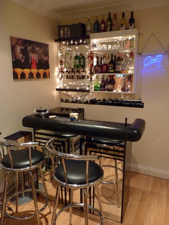 a stylish modern home bar with lots of wall mounted shelves covered with lights, a fridge on the floor, a mirror stand and tall black stools