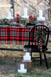 a rustic Christmas tablescape with a plaid tablecloth, evergreens, berries, bells and pillar candles on wood slices