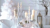 a chic white winter tablescape with a silver runner, bells, various candles and a large candle lantern