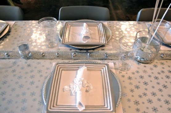 a white and silver winter tablescape with a snowflake tablecloth, bell table runner, silver chargers and plates
