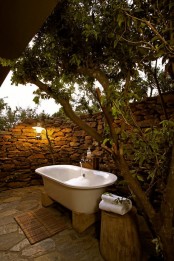 a rustic outdoor bathroom with a stone wall, a tub on stands, stumps as side tables and living trees