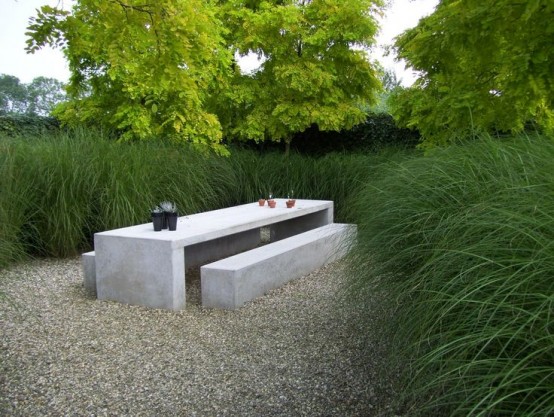a minimalist outdoor space with tall grass, a white concrete table and benches and mulch on the ground is a lovely space to have meals