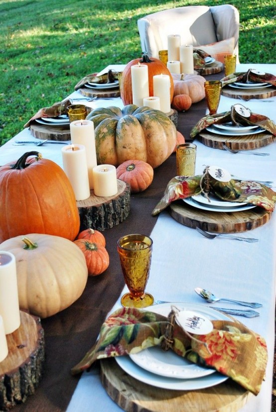 a modern rustic Thanksgiving table setting with a runner with wood slices, real pumpkins and pillar candles, colorful napkins and colored glasses