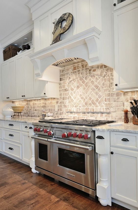 a white farmhouse kitchen with shaker style cabinets, black knobs, a whitewashed brick backsplash with a pattern and built in lights and stainless steel appliances