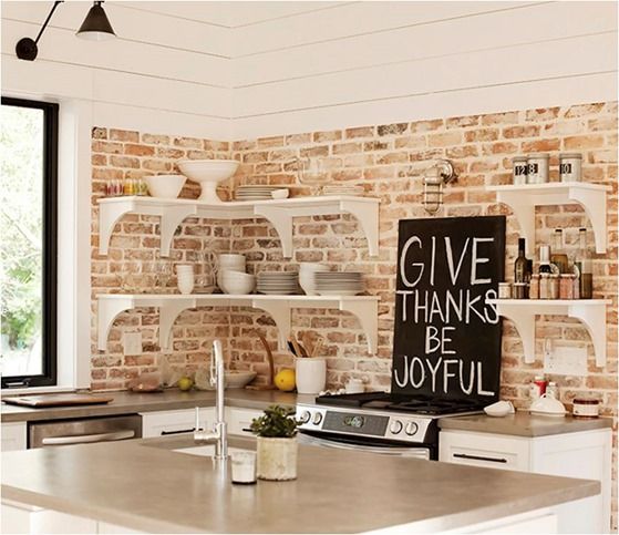 a white kitchen with shaker style cabinets and black handles, with open shelves, a whitewashed brick backsplash and a small kitchen island with a concrete countertop