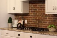 a chic kitchen with white cabinetry, grey countertops, a red brick backsplash, a vintage hood painted white is a welcoming space