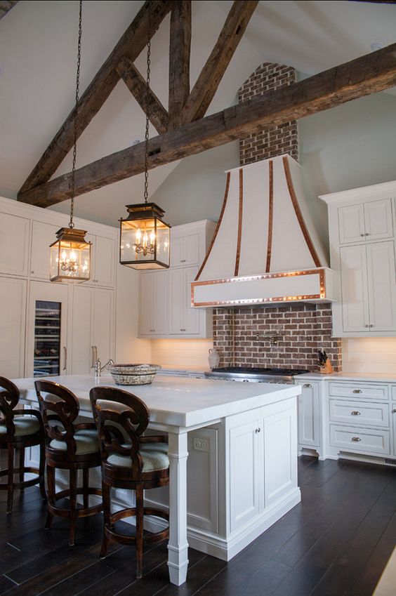a vintage white kitchen with shaker style cabinets, built in lights and a large hood with copper detailing, a red brick backsplash for a touch of color and more interest, pendant lamps and dark stained stools