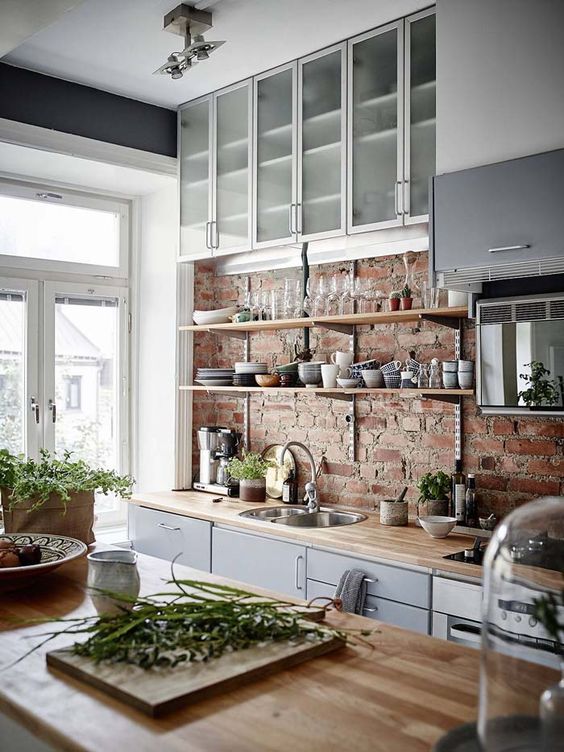a sleek grey Scandinavian kitchen with sleek butcherblock countertops, a rough red brick backsplash, frosted glass cabinets and lots of natural light incoming