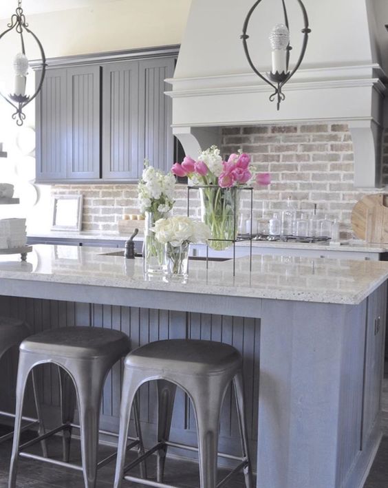 a farmhouse kitchen with grey beadboard cabinets, a brick backsplash and a white hood, white countertops, vintage pendant lamps