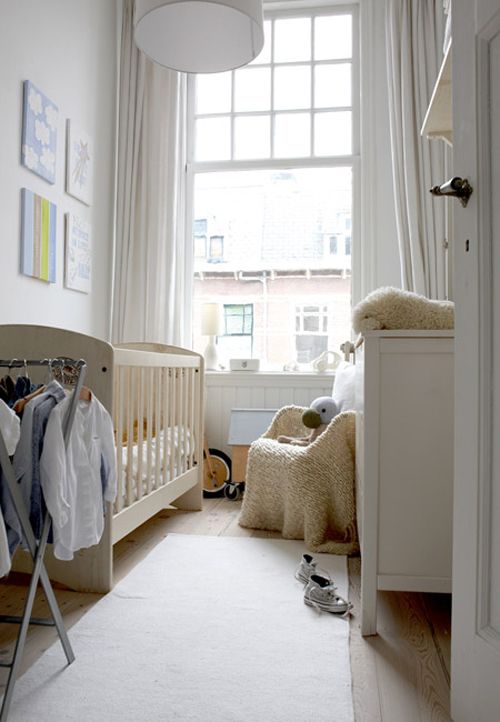 a tiny white nursery with a neutral crib, a white dresser, a neutral chair, a makeshift closet and some artwork and toys