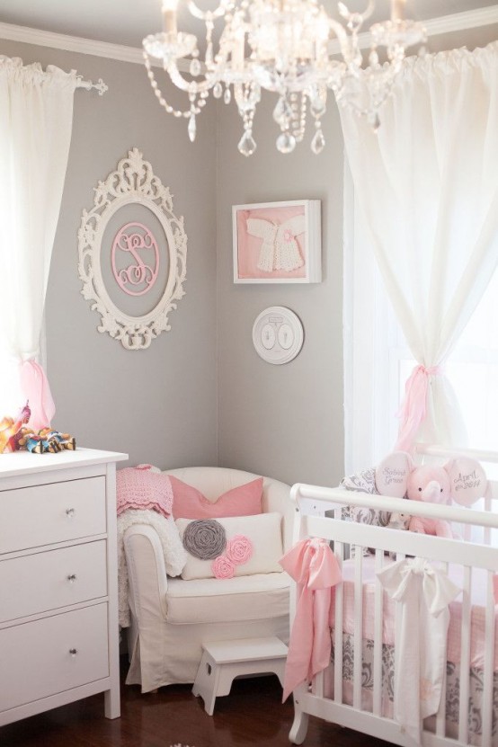 an elegant tiny nursery with grey walls, pink and white artwork, a white crib and a dresser, a white chair and some pillows, a chic vintage chandelier