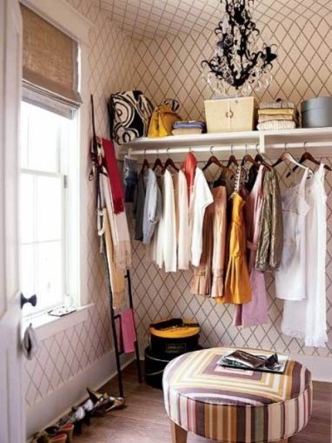 a pretty girlish walk in closet with printed wallpaper, an open storage unit, a black crystal chandelier, a colorful geometric ottoman and a ladder